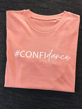 Load image into Gallery viewer, A folded peach-colored t-shirt with the text '#CONFIDENCE' printed vertically in white script, laid flat on a dark textured background.